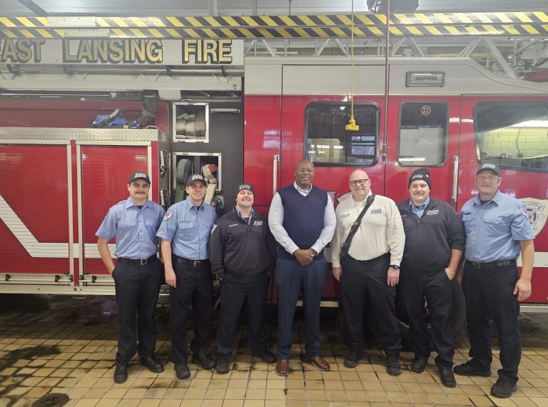 Chuck Grigsby stands in front of a fire engine with six firefighters in casual uniform.