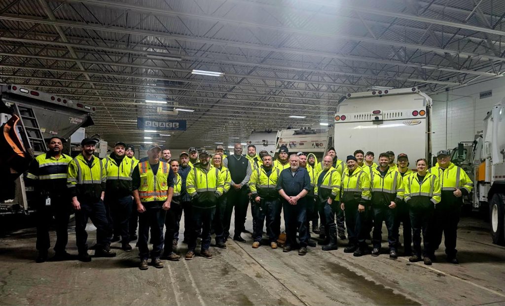 Several dozen workers in high-visibility jackets stand in an industrial garage, with Chuck Grigsby in the center. A line of garbage trucks and salt trucks can be seen in the background.
