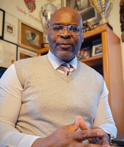 A pensive portrait photo of Chuck Grigsby. He is wearing a cream-colored vest and time and had his hands folded in front of him.