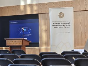 A tall banner standing in the front of a room reads, Commission to Study the Potential Creation of a National Museum of Asian Pacific American History and Culture. Beside it is a wood podium with a gold plaque that reads, Michigan State University. Rows of folding chairs are in the foreground facing the podium and banner.