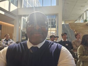 A selfie of Chuck Grigsby smiling in a blue vest and tie. He is seated in an audience of primarily Asian Pacific Americans who are listening intently to a speaker at the podium.