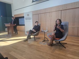 A man and a woman are seated in stylish chairs in the front of a room. The woman holds a microphone and gestures while speaking about the National APA Museum.